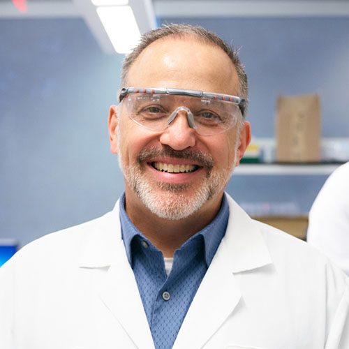 Man wearing safety glasses and white lab coat in a lab setting.